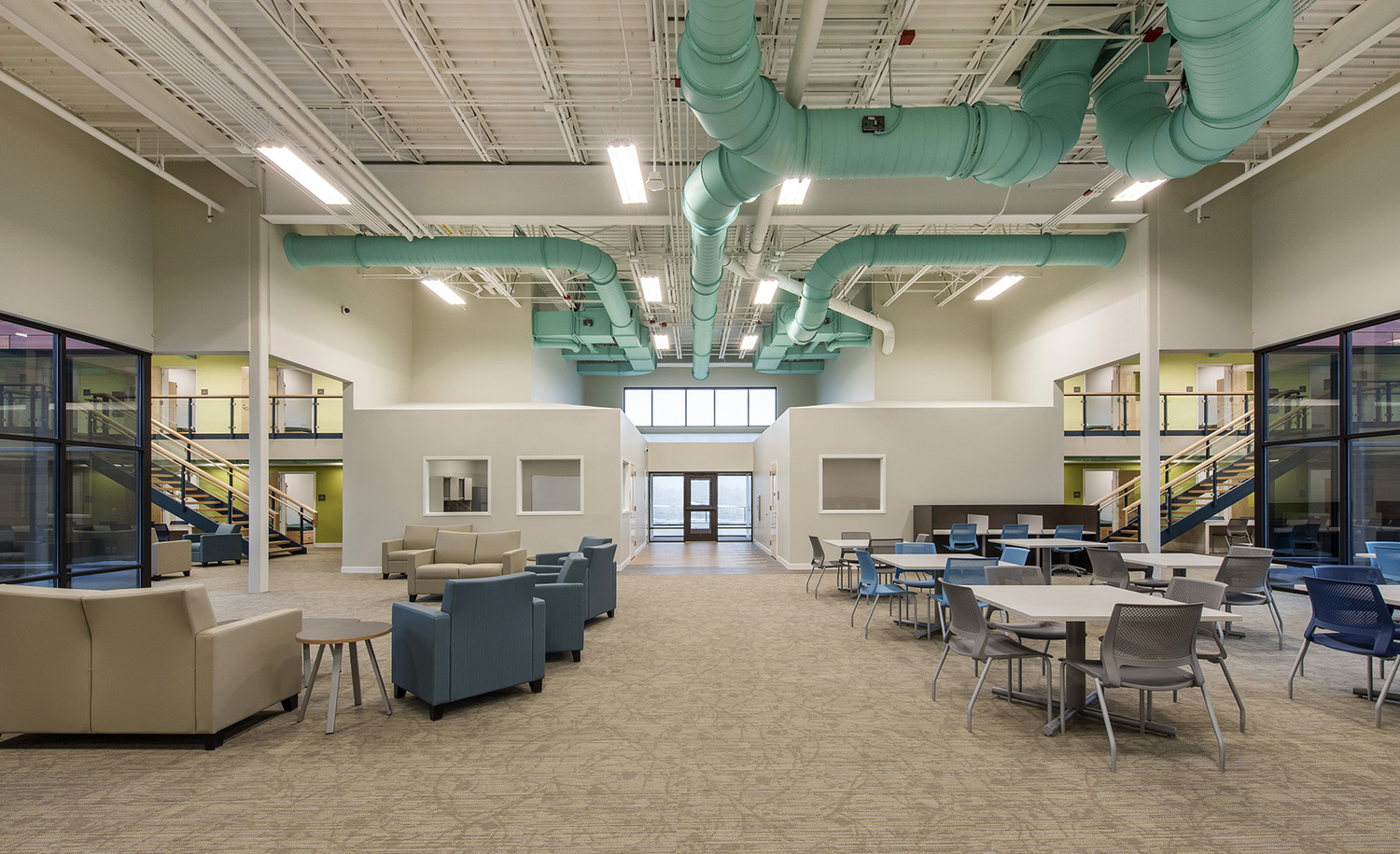 Brightly painted duct work provides a colorful accent to the central great room. To the left, upholstered chairs and couches create a lounge space for residents. To the right, tables and chairs provide room for activities.